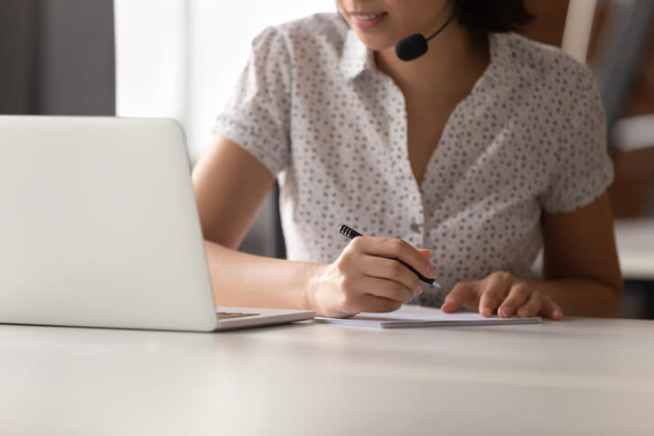 woman on phone headset looking at laptop and taking notes